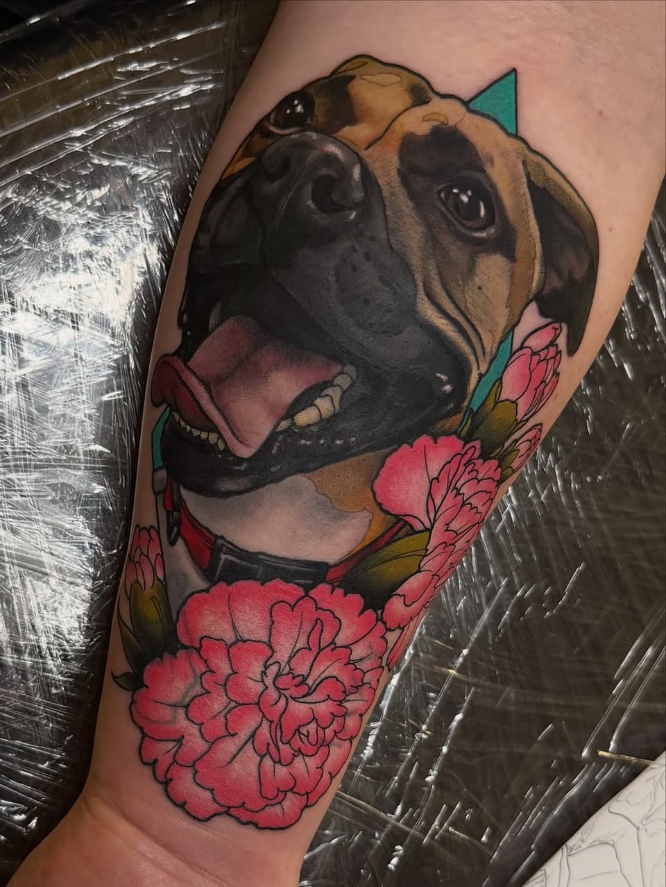Colorful dog head tattoo with open mouth on upper arm, surrounded by pink flowers and teal accents on the wearer's skin.