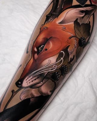 A vibrant red fox head with berries and botanical elements tattooed on an arm, rendered in orange, brown, and black colors.