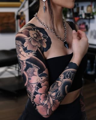 Woman displaying a full arm sleeve tattoo featuring birds, flowers, and botanical elements in black ink with shading on her shoulder and forearm.