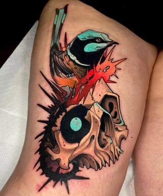 Colorful bird with turquoise head perched on a skull with red spikes and black spines, tattooed on upper thigh.