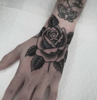 Large rose with leaves and stem tattooed on the back of a hand and forearm in black and gray ink.