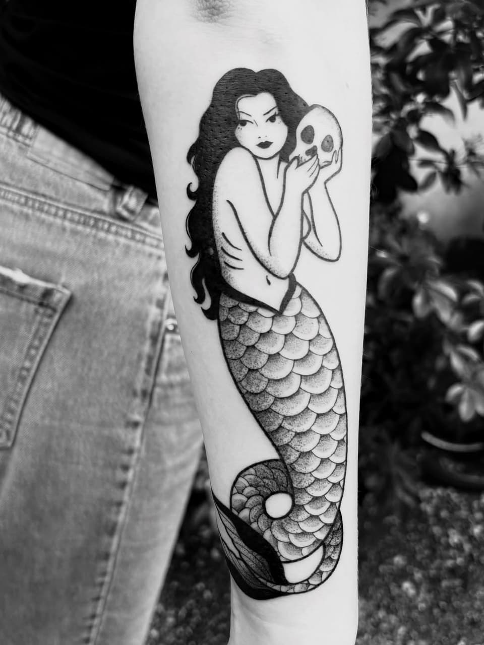 Mermaid holding a skull tattoo on the upper arm in black ink, featuring long dark hair, scaled tail, and detailed linework.