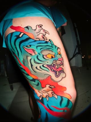 Vibrant turquoise tiger head with red and orange flowing ribbons on upper arm, featuring pink mouth and yellow eyes with detailed line work.
