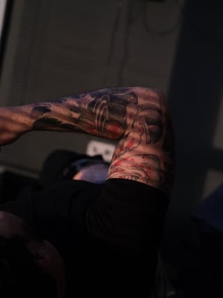 Detailed skull tattoo on forearm featuring red and black ink with ornamental flourishes and facial details, displayed on muscular arm.