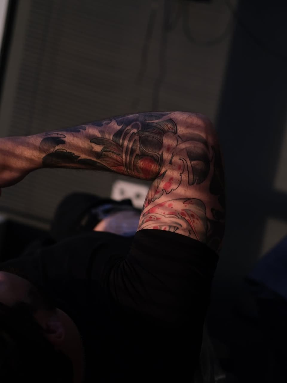Detailed skull tattoo on forearm featuring red and black ink with ornamental flourishes and facial details, displayed on muscular arm.