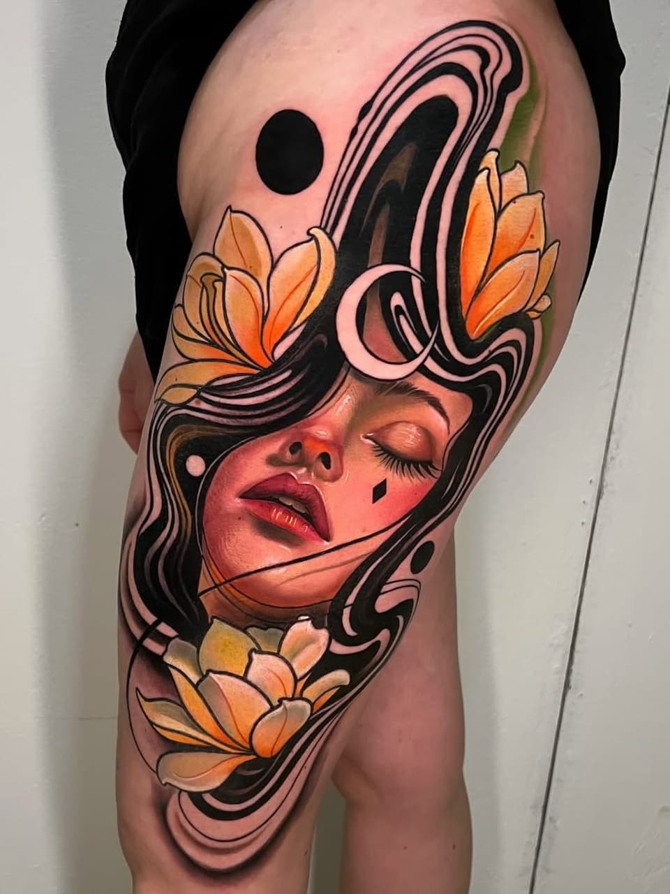 Woman's thigh tattoo featuring a female face with closed eyes, orange flowers, swirling black linework, and celestial elements in warm peachy and orange tones.