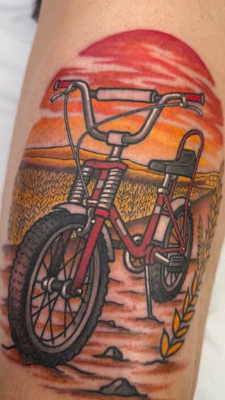 Red bicycle with drop handlebars positioned in a desert landscape with golden wheat fields and warm sunset sky, tattooed on an arm.