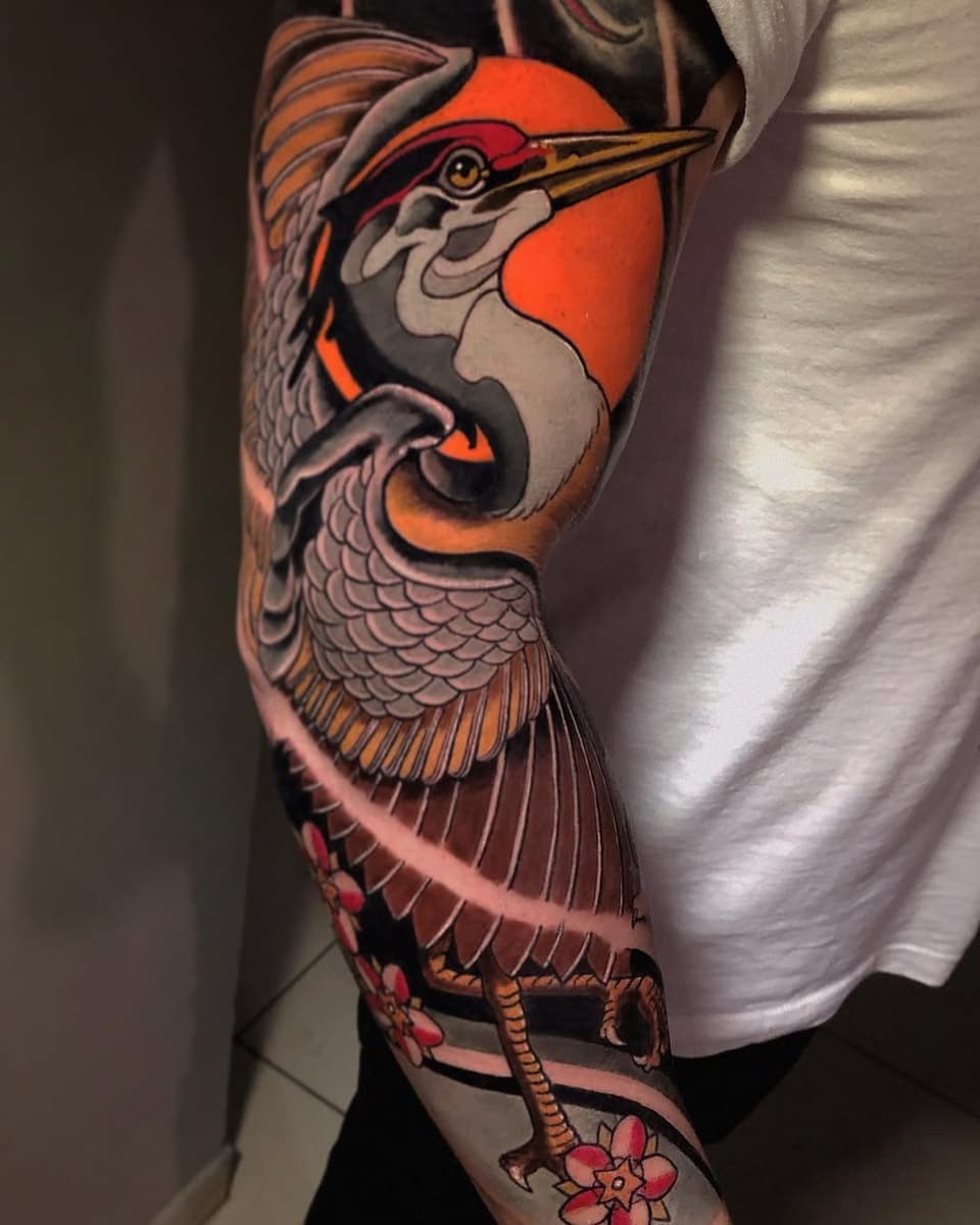 Colorful heron tattoo on arm featuring orange, red, and grey tones with detailed feathers and floral elements at the wrist.