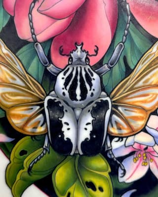 Colorful beetle tattoo with white and black body, golden wings, surrounded by pink roses and green leaves on the upper arm.