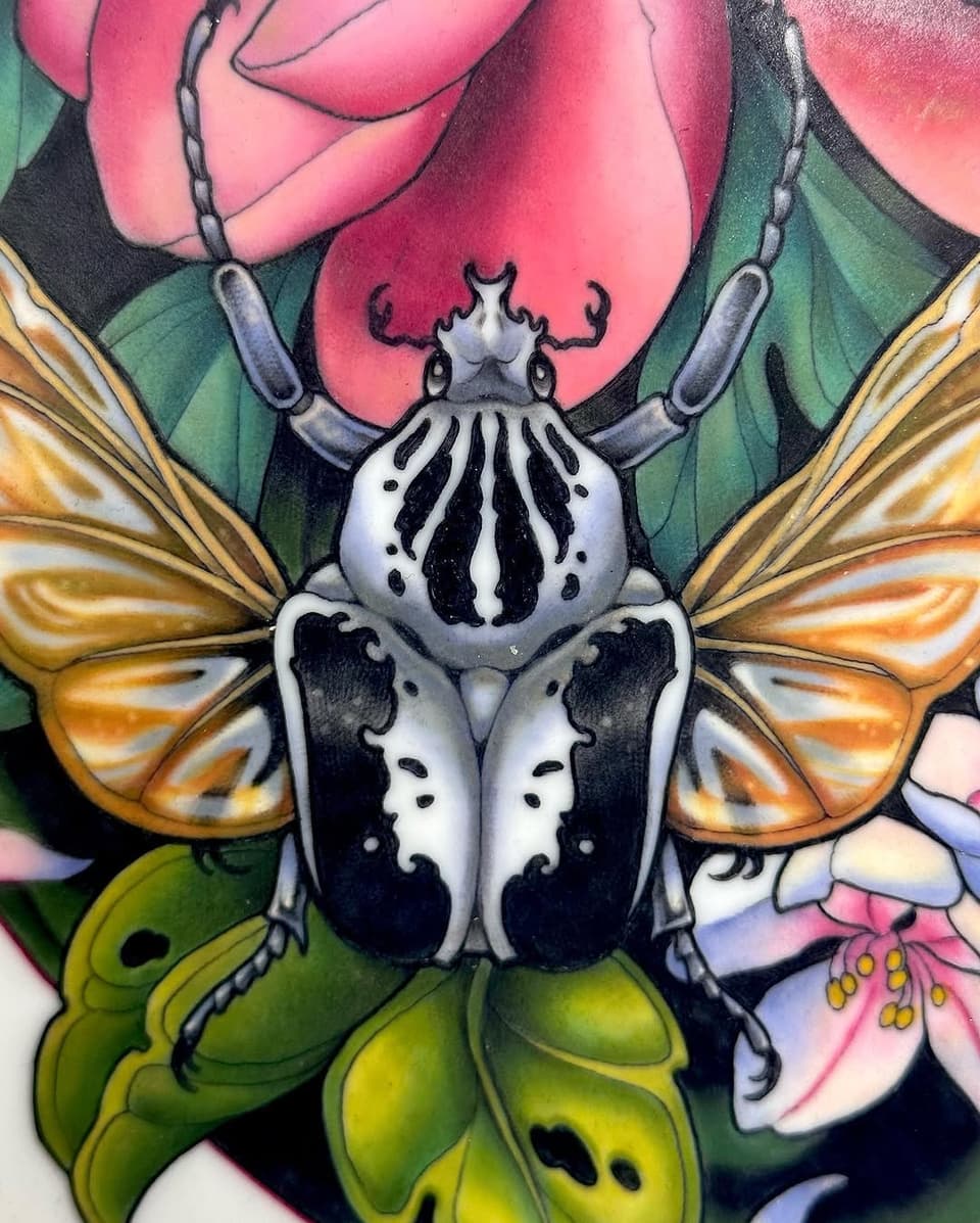 Colorful beetle tattoo with white and black body, golden wings, surrounded by pink roses and green leaves on the upper arm.