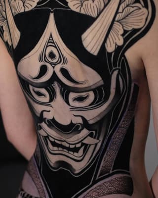 Large demon mask tattoo on thigh featuring intricate facial details, menacing expression, and decorative floral elements above, rendered in black ink with shading.