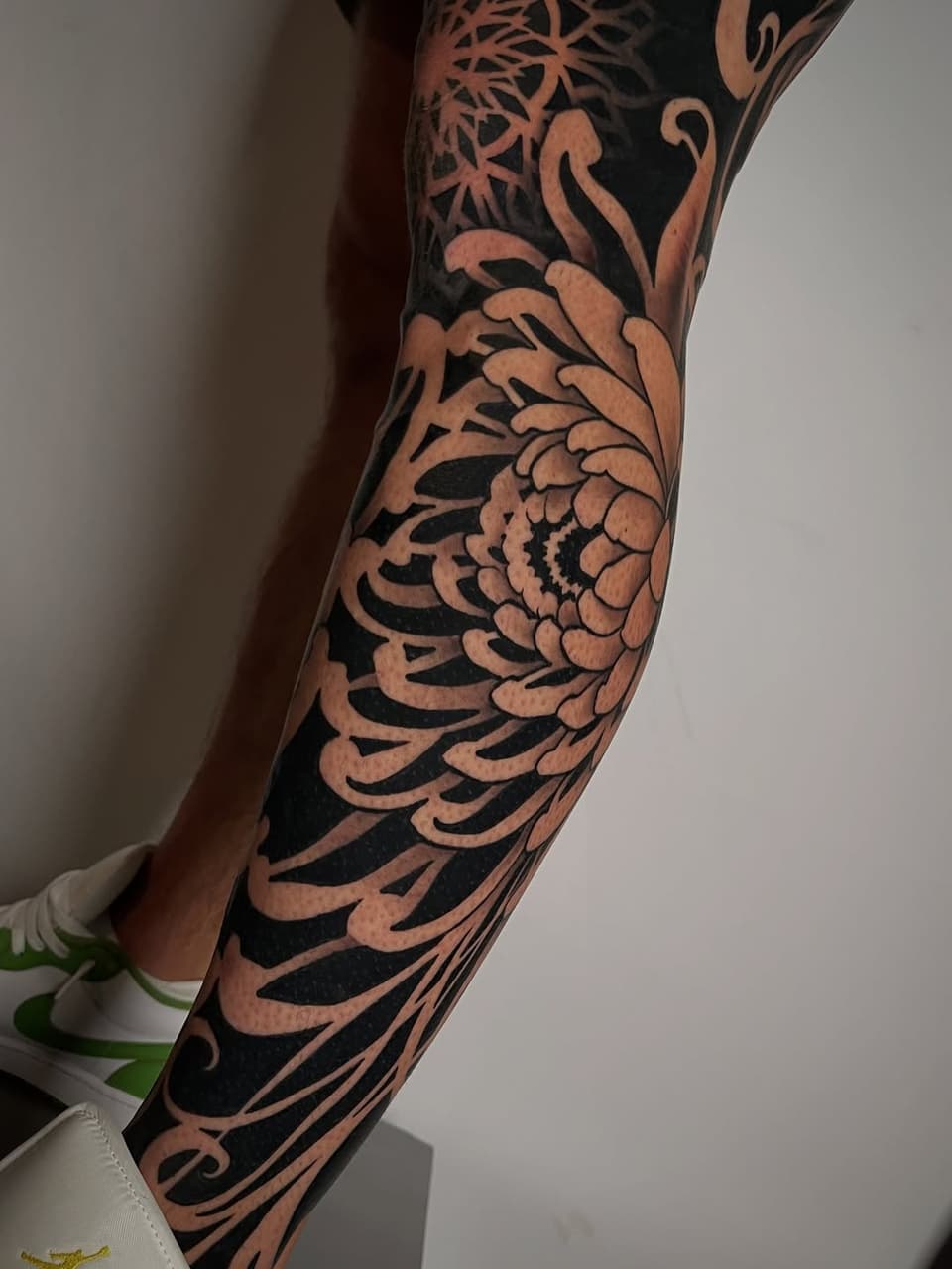 Detailed arm sleeve tattoo featuring a large chrysanthemum flower with ornamental leaves and curved linework in black ink on tan skin.