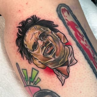 Colorful tattoo on upper arm depicting a screaming face with black hair, stitched features, and red and green accents surrounding the design.