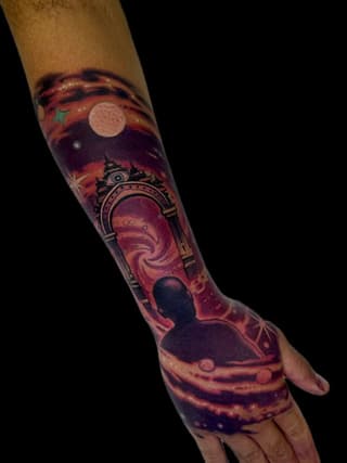 Colorful tattoo on forearm and hand featuring a portal with swirling interior, celestial elements including moons and stars, and a silhouetted figure against warm orange and purple tones.