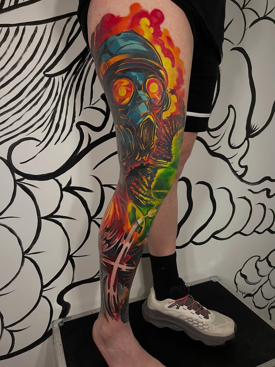 Full-leg sleeve tattoo featuring a skull with glowing eyes and gas mask, surrounded by vibrant red, orange, yellow, and green flames and abstract elements.