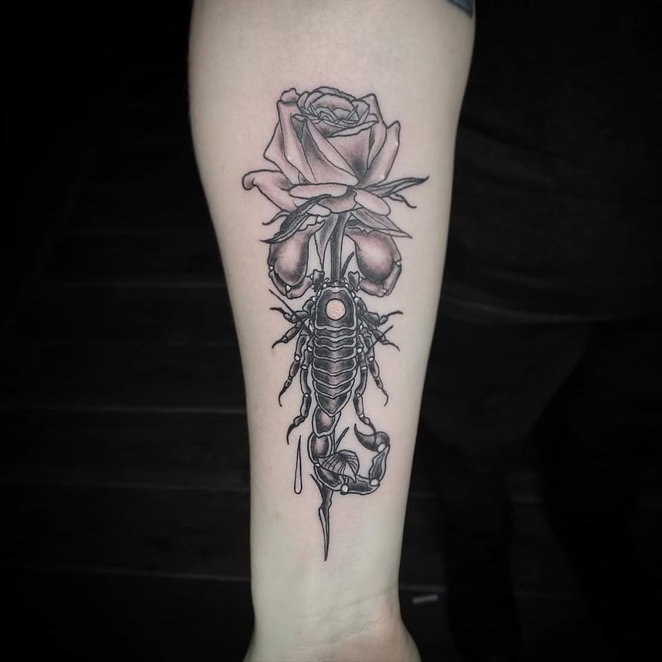 Vertical tattoo on pale forearm featuring a rose at top, bell-shaped flowers in middle, detailed insect with spotted center, and dripping element below in black ink.