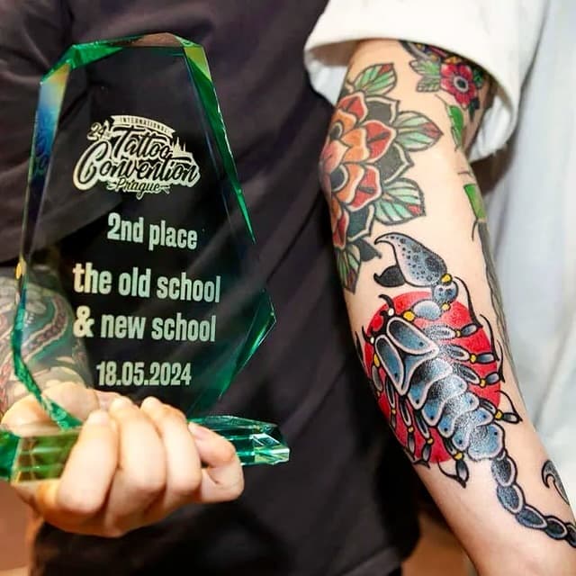 Colorful arm tattoo featuring red flowers, green leaves, and birds displayed next to a green award trophy dated 18.05.2024.