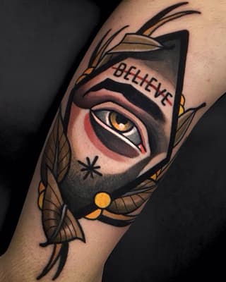 Colorful tattoo on upper arm featuring a geometric cat face with golden eyes, feathered wings, and "BELIEVE" text on the forehead.