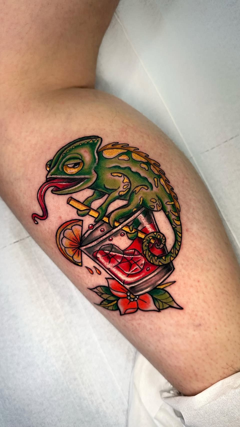 Green chameleon with extended tongue holding an orange slice above a red mouth and flower design, tattooed on the upper leg.