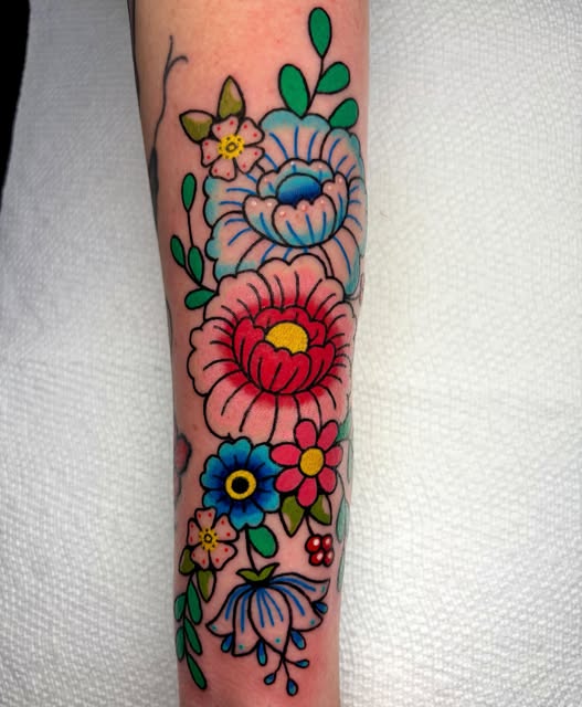 Colorful floral tattoo on forearm featuring stacked flowers in pink, red, blue, and yellow with green leaves and small decorative elements.