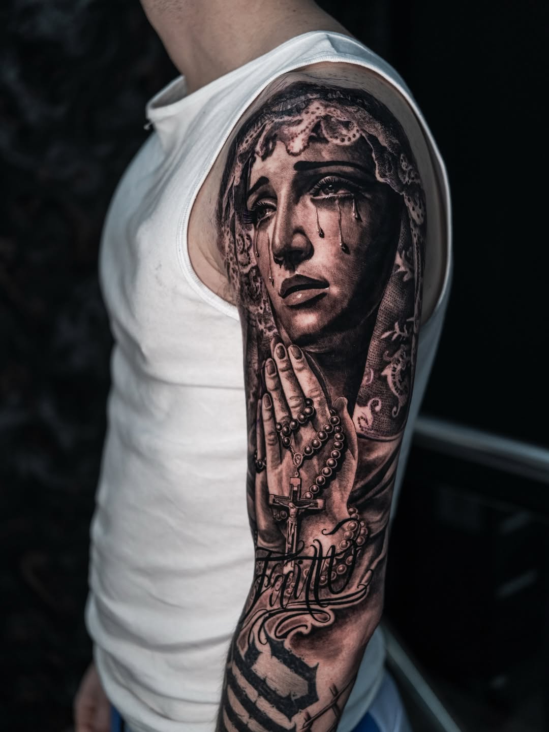 Photorealistic portrait of a woman with lace veil and rosary beads, rendered in black and grey on upper arm.