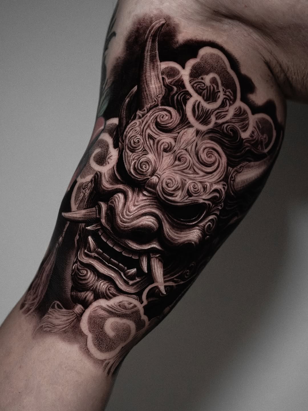 Black and grey ornamental demon mask tattoo with intricate swirling details on the upper arm.