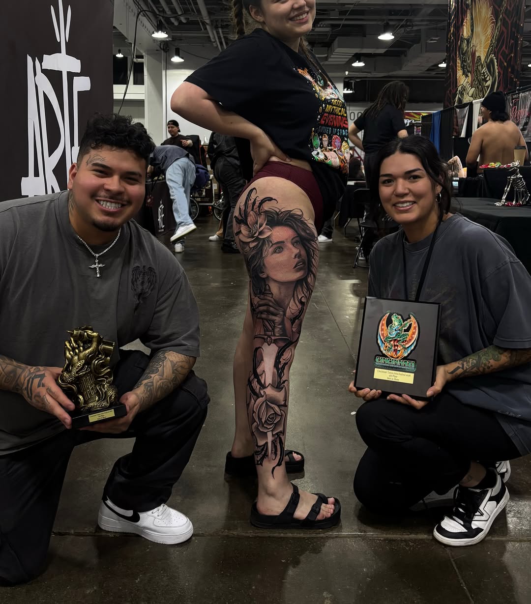 Black and grey portrait tattoo of a woman's face with flowers on the thigh, displayed by a tattoo artist at a convention.