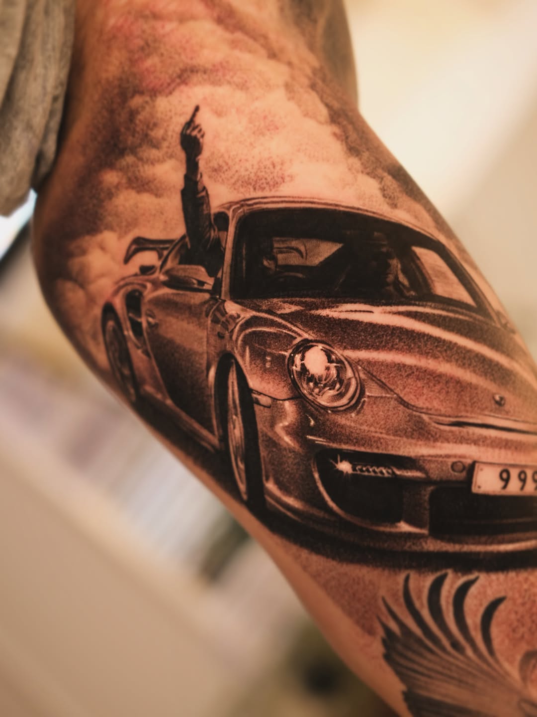 Black and grey realistic car tattoo on arm showing vintage Porsche with open door and figure, detailed shading.