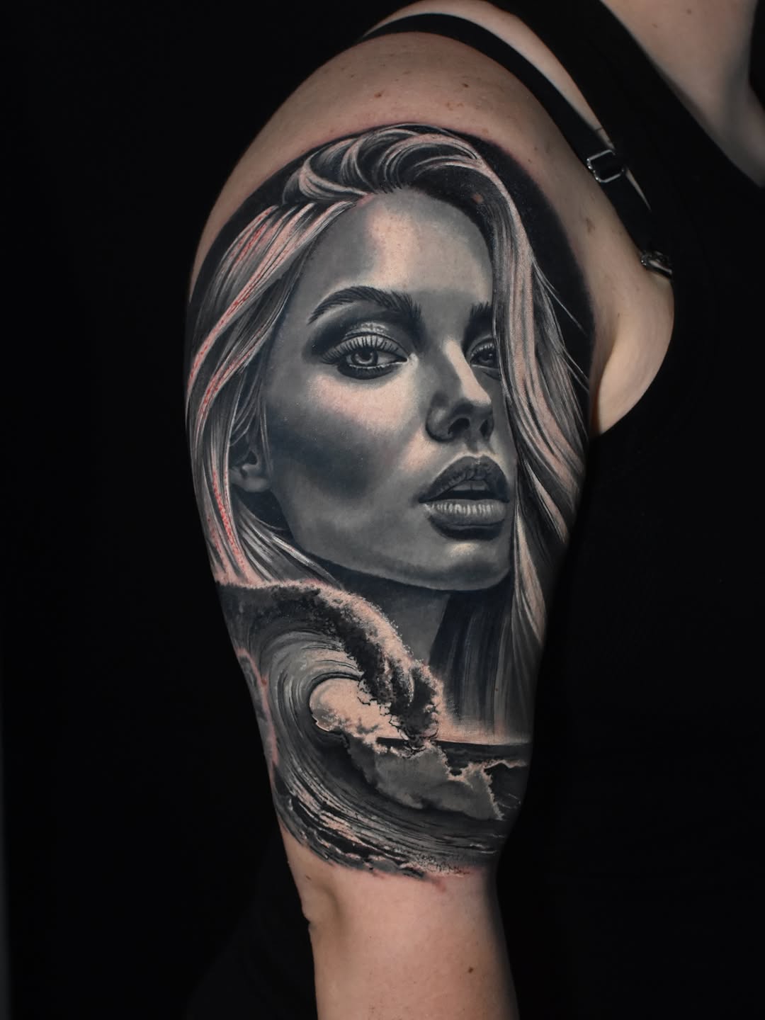 Black and grey tattoo of a woman's face with flowing hair above an ocean wave, on upper arm shoulder.