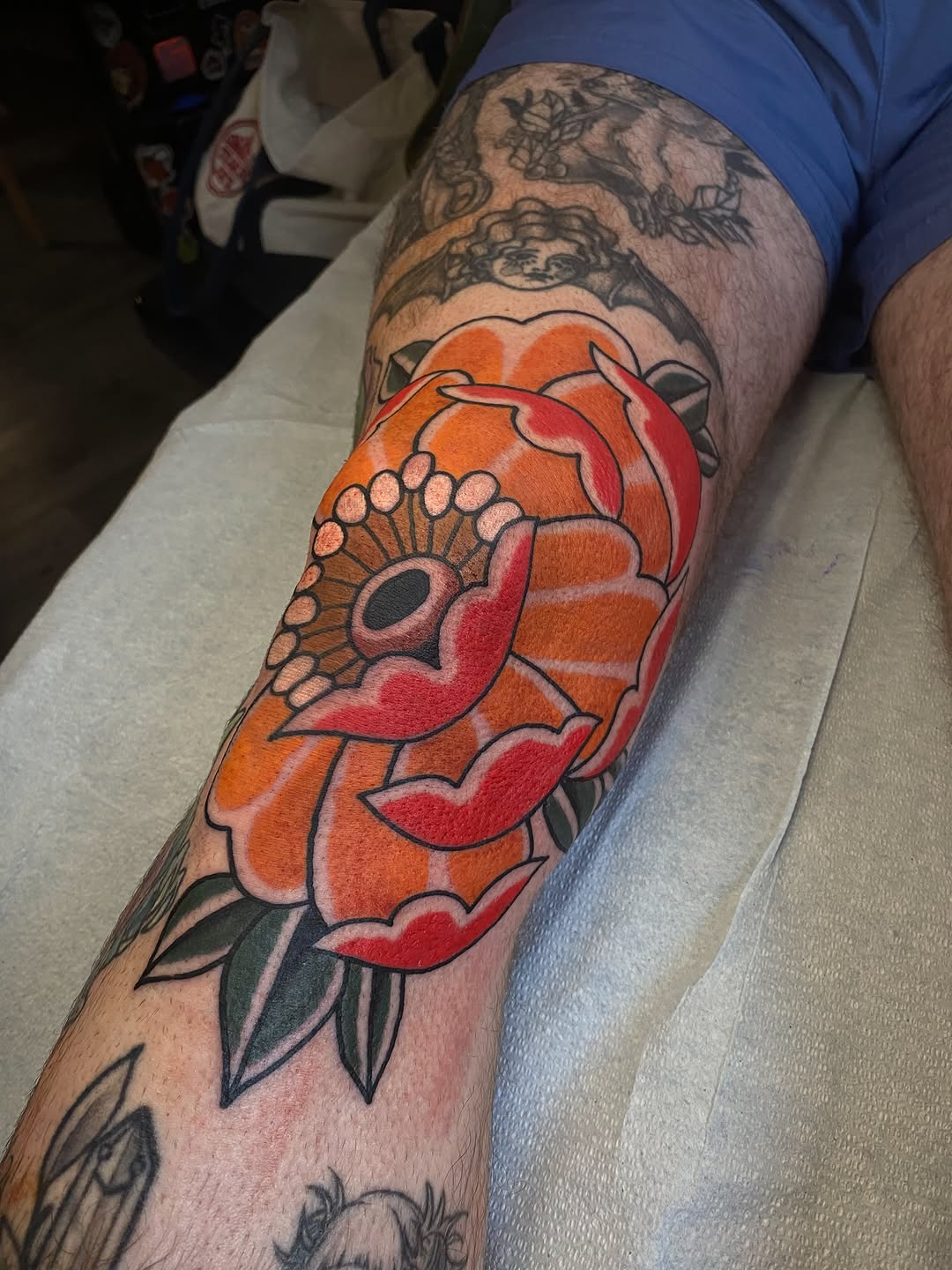 American Traditional rose tattoo with orange and red petals, bold black outlines, and green leaves on the forearm.