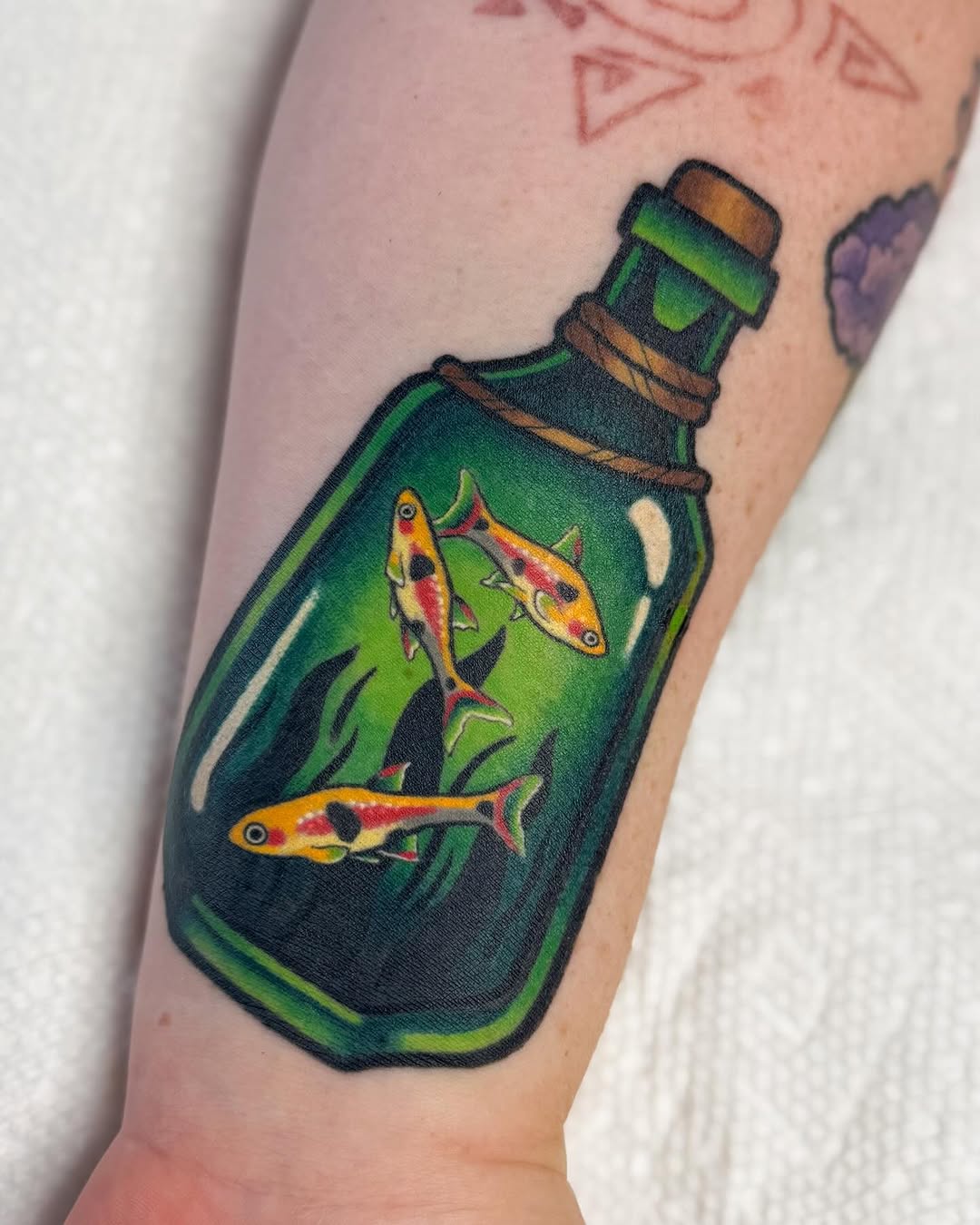 A green glass bottle containing three colorful fish with yellow, red, and black markings, tattooed on the forearm.