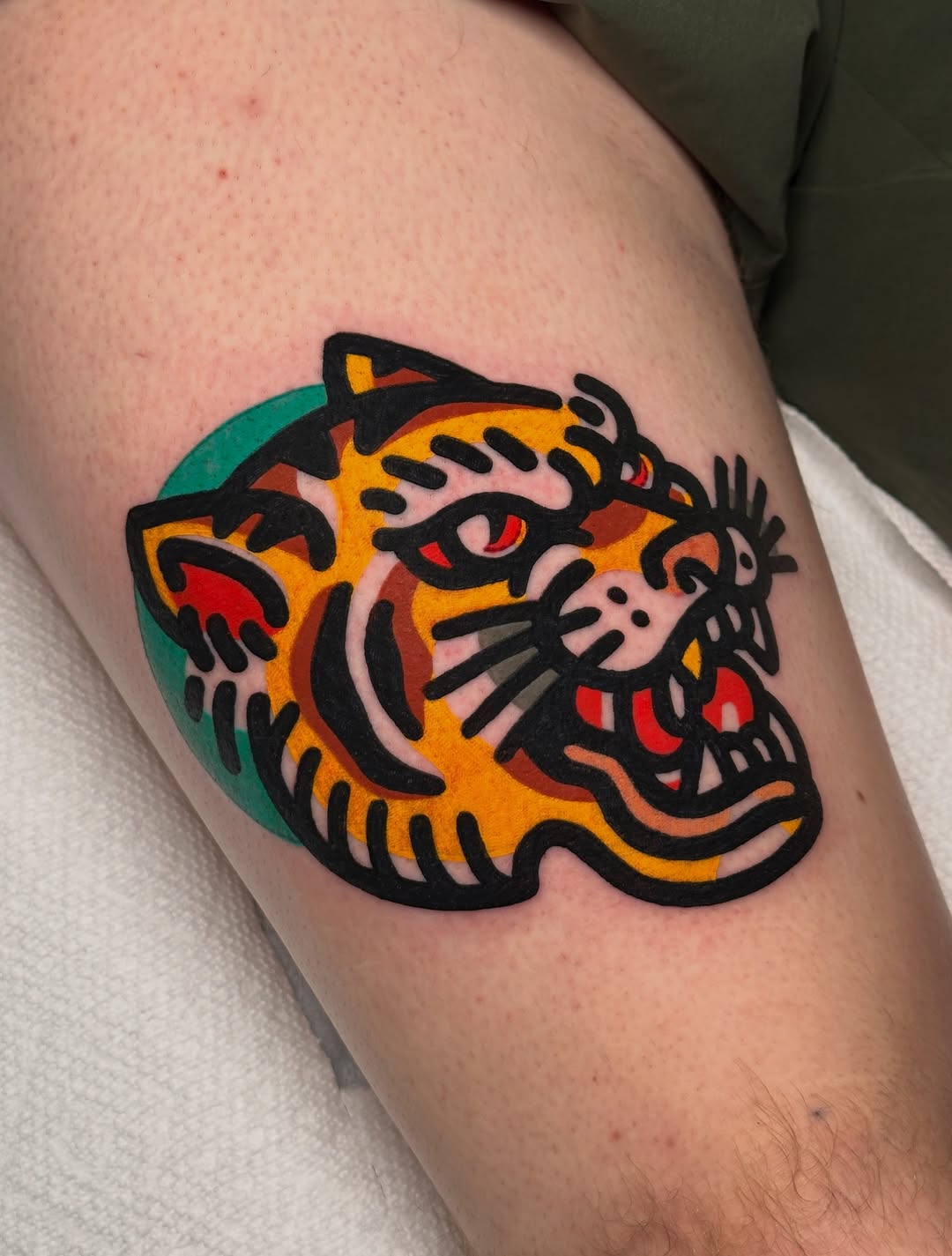 Colorful tiger head with bold black outlines, orange and yellow fill, red eyes and accents, green background circle on upper arm.