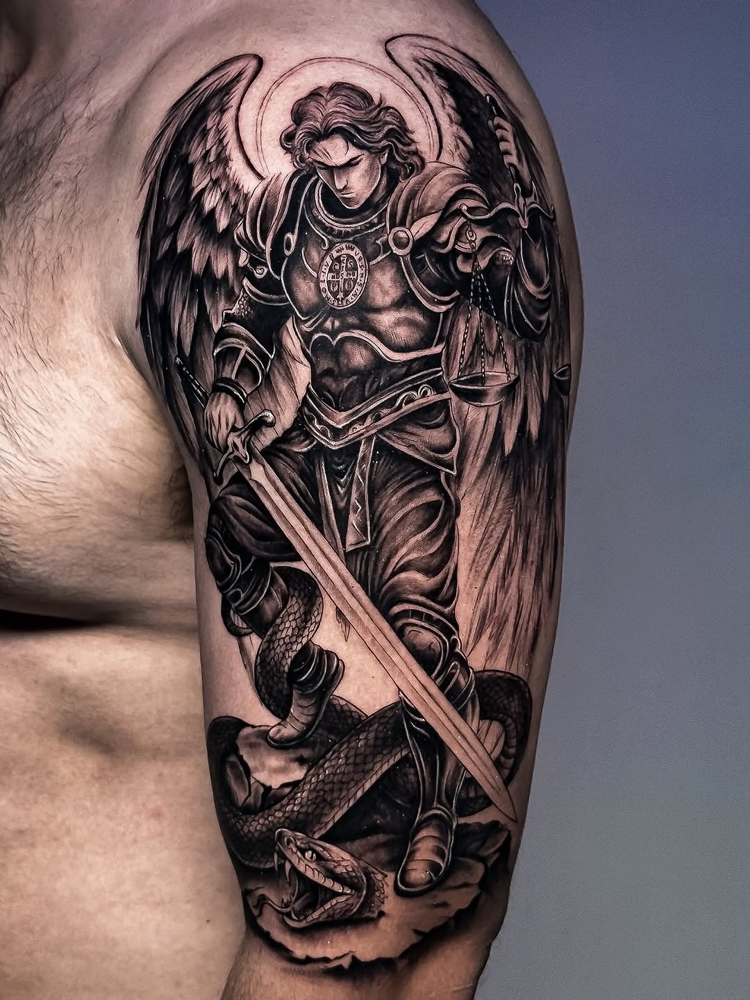 Saint Michael archangel with wings, armor, scales of justice, and sword defeating demon serpent on upper arm.
