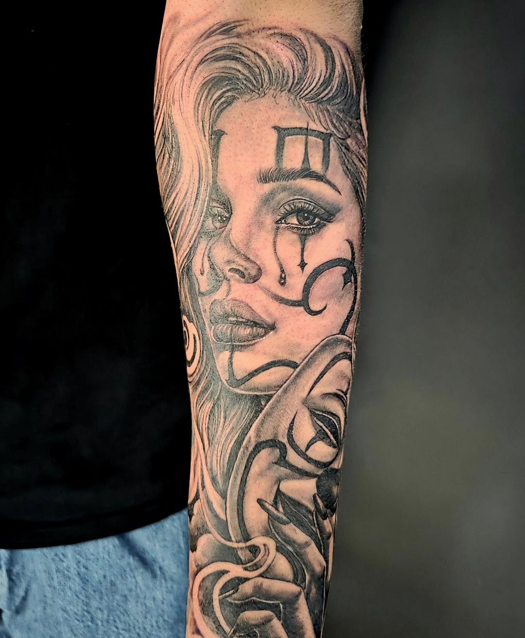Black and grey portrait tattoo of a woman with wavy hair, tears, geometric forehead symbol, and ornamental neck design on forearm.