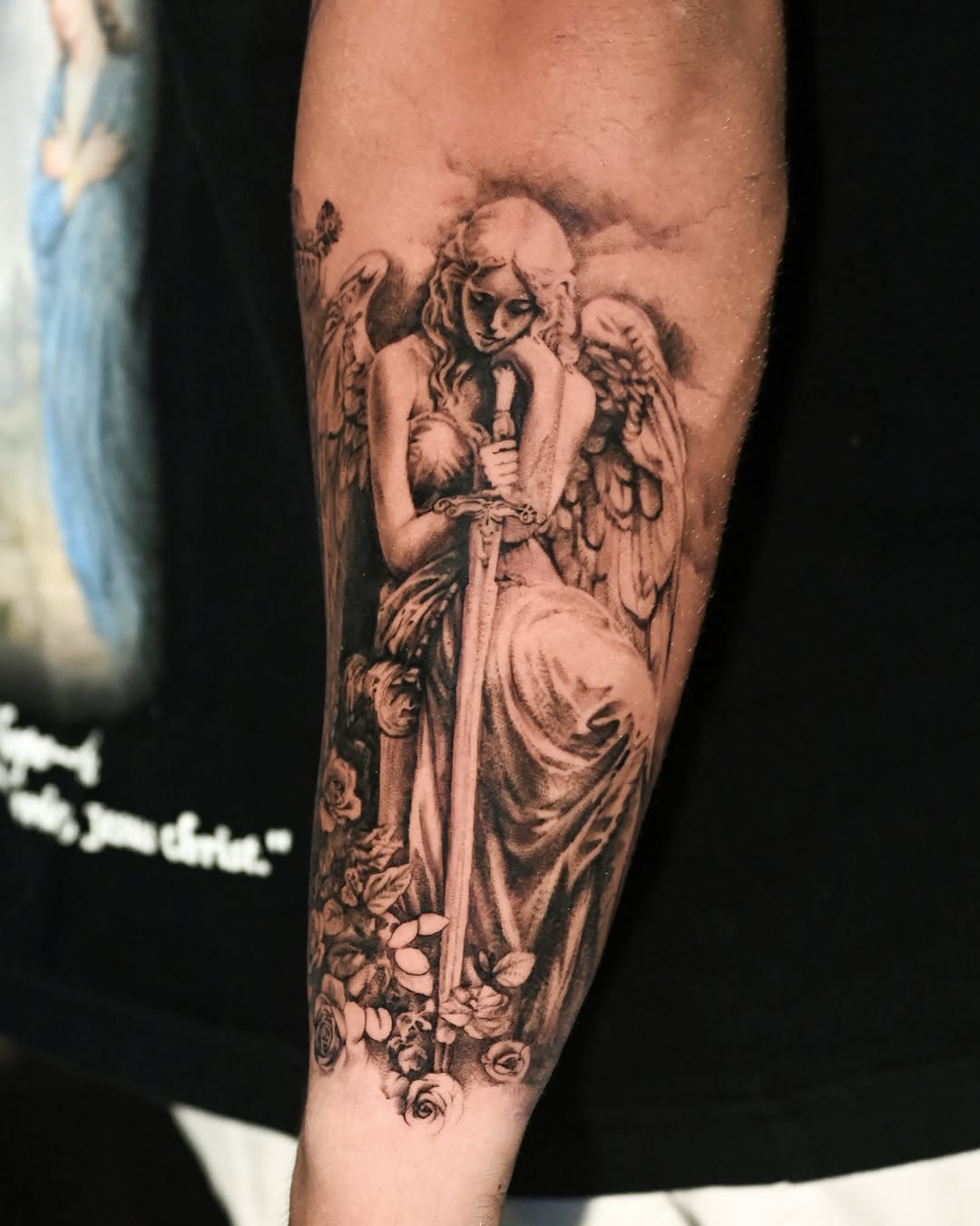 Black and grey angel figure with wings and flowing robes surrounded by roses, tattooed on forearm with realistic shading.