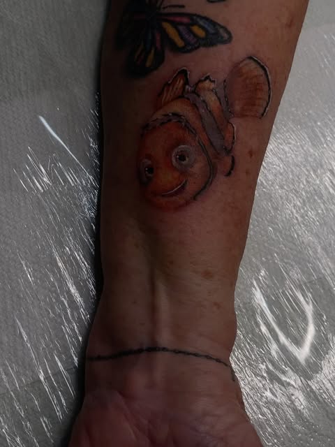 Nemo the clownfish tattoo on forearm with orange and white coloring, accompanied by a small butterfly above in black and yellow.