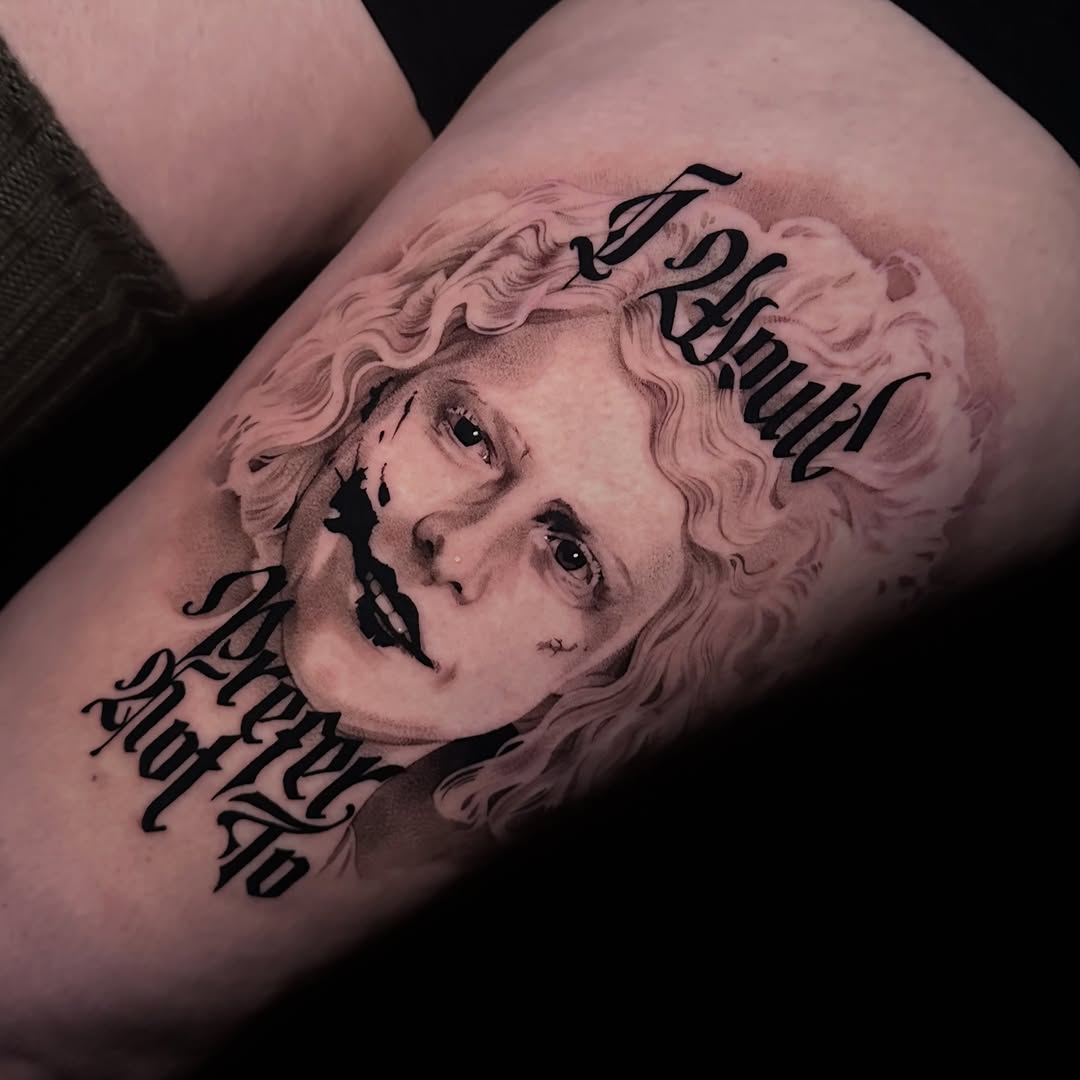 Portrait of a curly-haired woman with distressed lips and ornate blackletter script around it, on a thigh, in shades of black and grey.