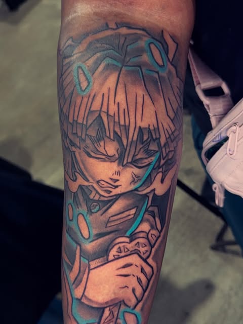 Anime character tattoo on forearm featuring a young person with spiky hair and teal accents, holding objects, rendered in orange and blue tones.