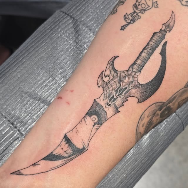 Ornate fantasy axe tattoo with wrapped handle and decorative blade on forearm, accompanied by smaller mystical symbols above.