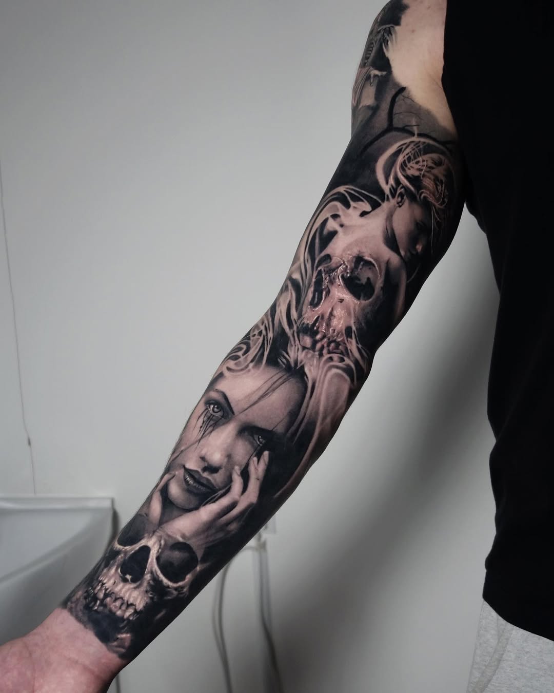 Full-sleeve monochrome portrait tattoo featuring skull and human faces with ornamental flourishes on the arm.