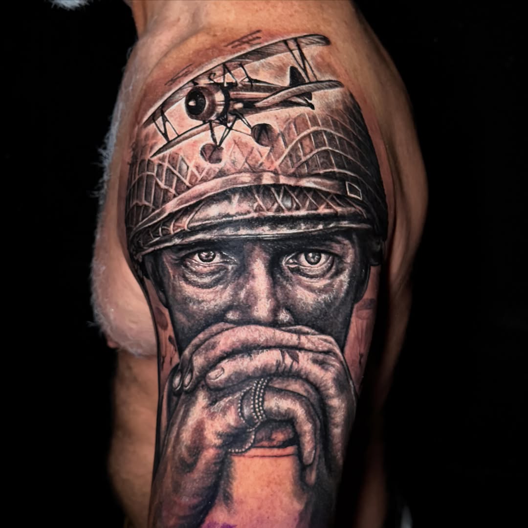 Black and grey portrait tattoo of a bearded man wearing a headwrap with a biplane above, on upper arm.