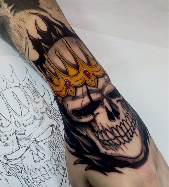 Skull tattoo with golden ornate crown featuring red jewels on the inner arm, with black shading and line work details.