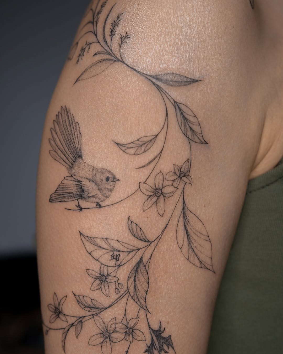 Delicate line-work botanical tattoo featuring a small bird perched among flowering branches and feathered leaf elements on the thigh.